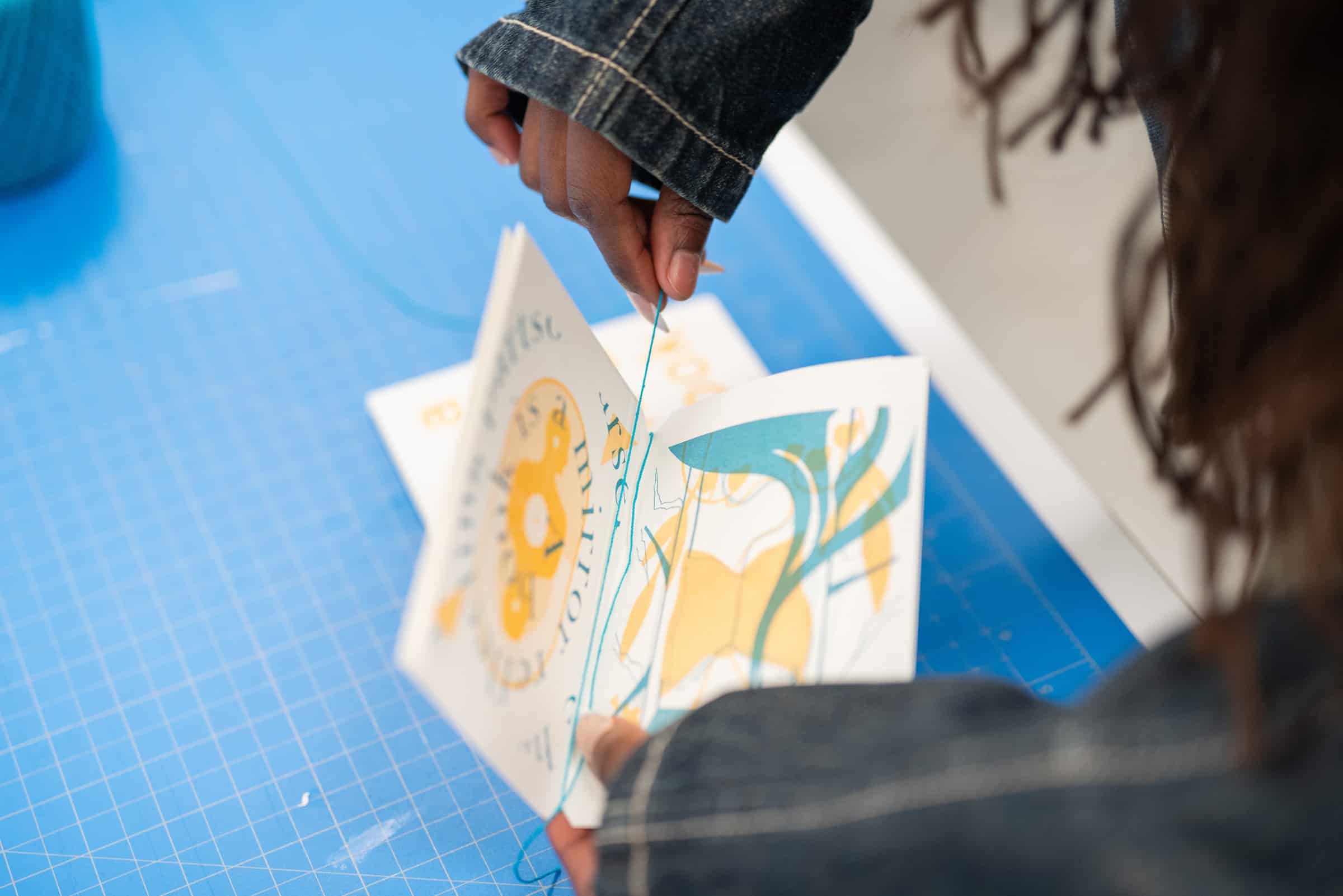 Close-up of hands stitching pages of a printed booklet together with blue thread.
