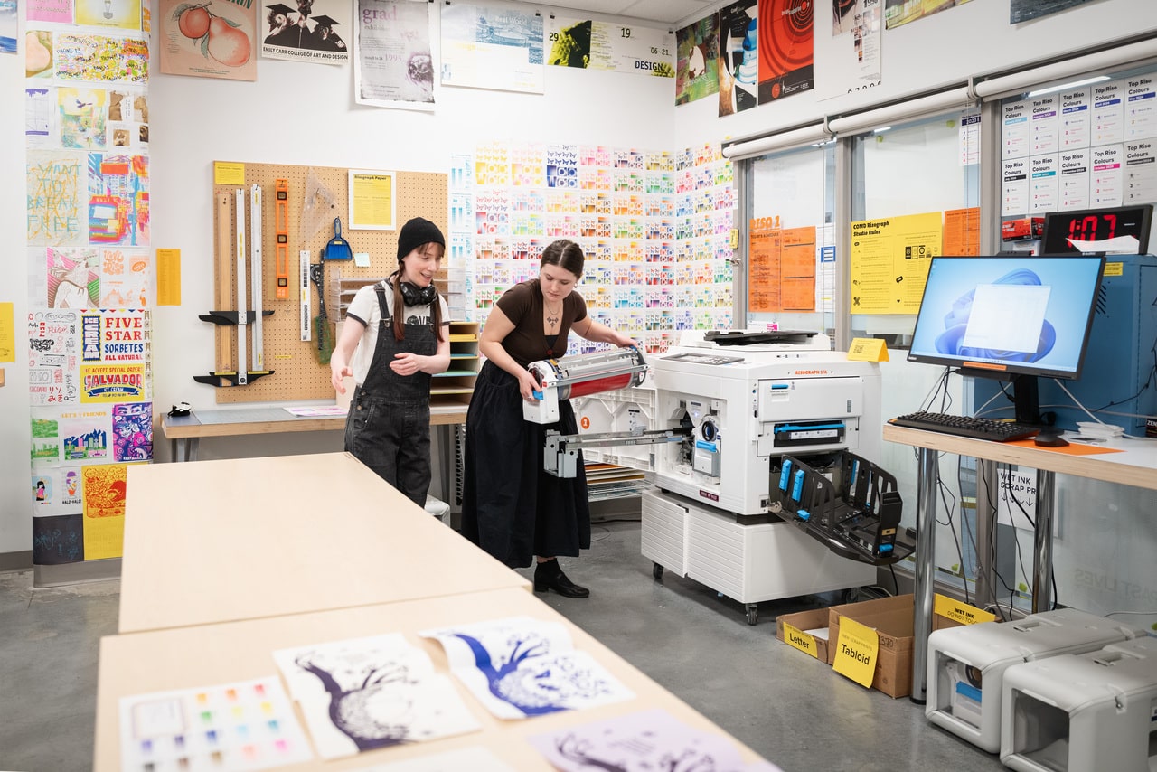 Two people operate a Risograph printer in a studio filled with colourful printed posters on the walls. Freshly printed artwork is laid out on a nearby table.