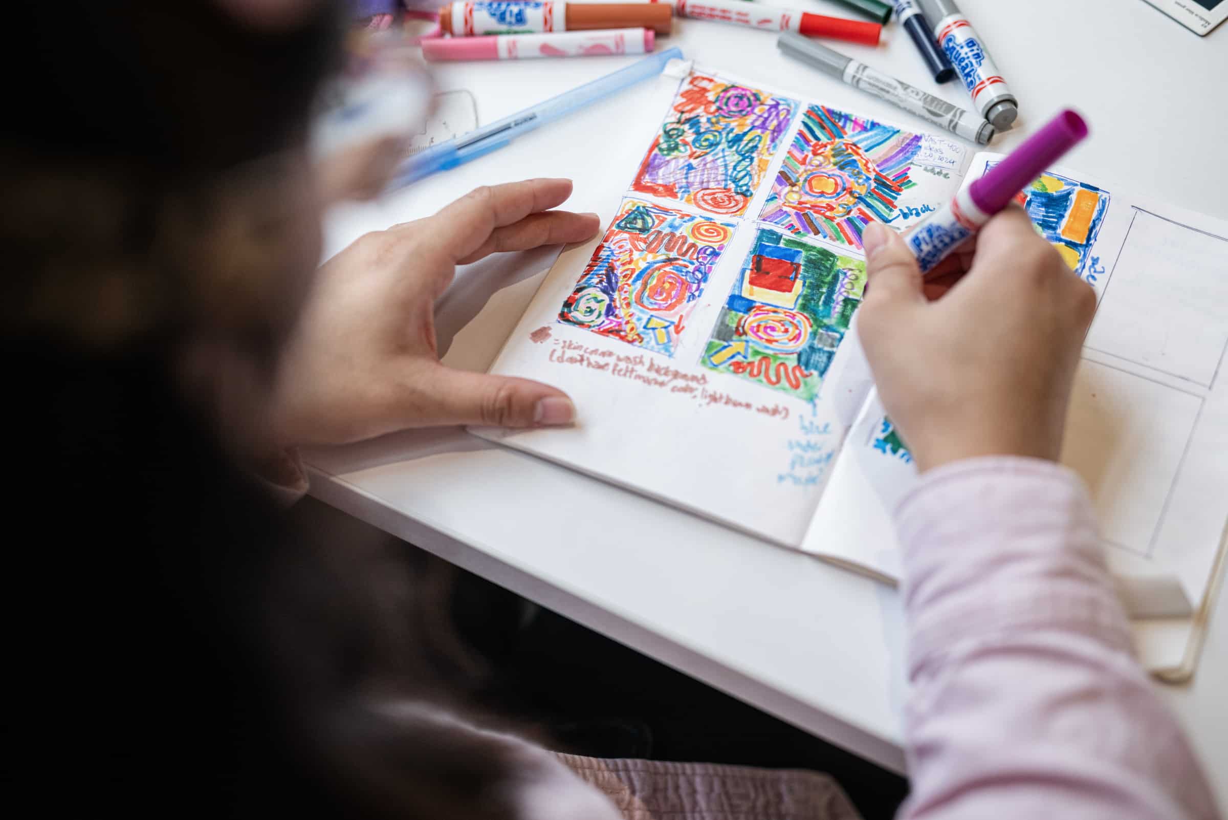 Close-up of a person drawing colorful abstract sketches with markers in a notebook.