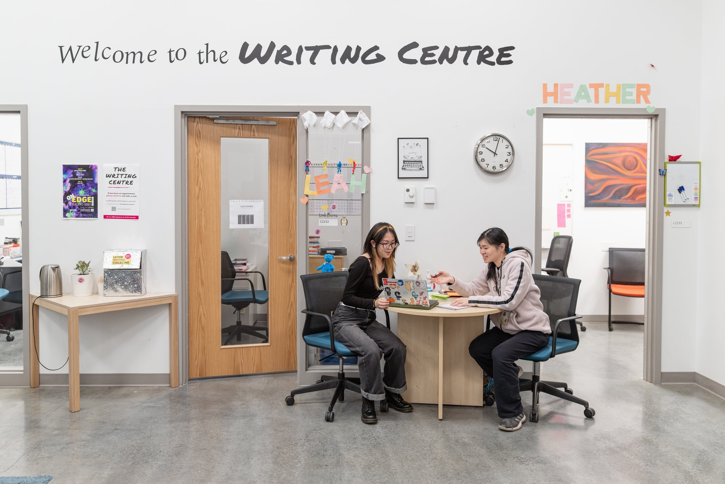 Two people sit at a small table in the Writing Centre, working together on a laptop. The wall behind them reads “Welcome to the Writing Centre” with bright name signs, posters, and a clock.