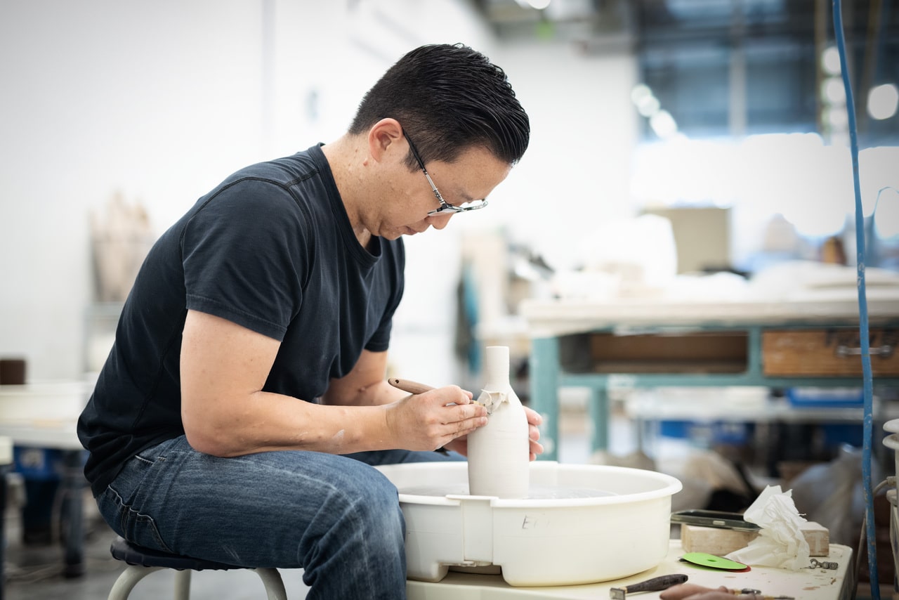 Person wearing glasses shapes a clay vessel on a pottery wheel, using a tool to refine the surface. The ceramics studio is visible in the background.
