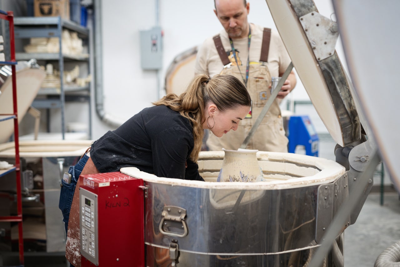 Person leans over an open kiln, carefully placing or inspecting a ceramic piece inside. Another person in overalls stands behind them, observing the process in the ceramics studio.