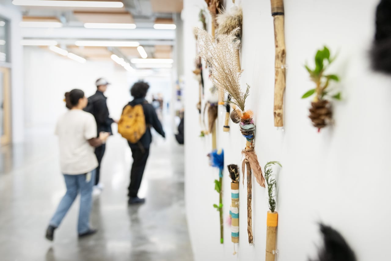 Mixed-media objects made from natural materials, feathers and colourful threads are mounted on a white wall in a hallway. In the background, several people walk past the installation, slightly out of focus.