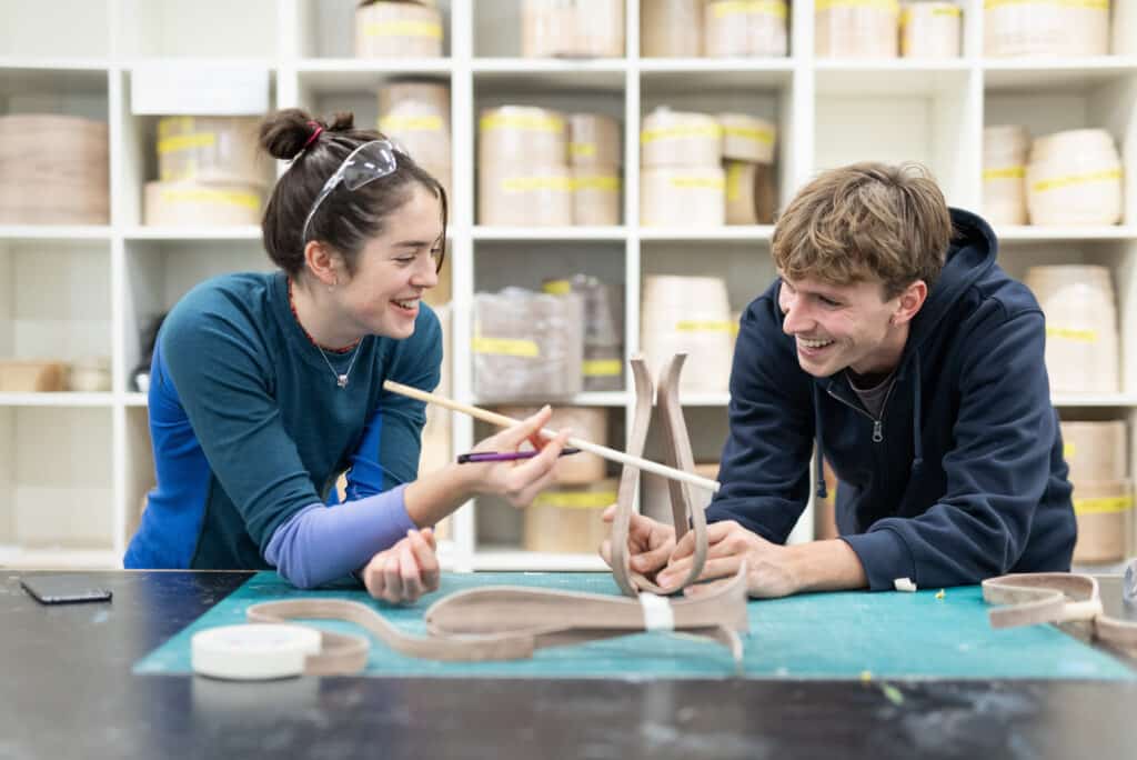 Two students laugh and collaborate while assembling a bentwood structure in a workshop, surrounded by shelves of materials.