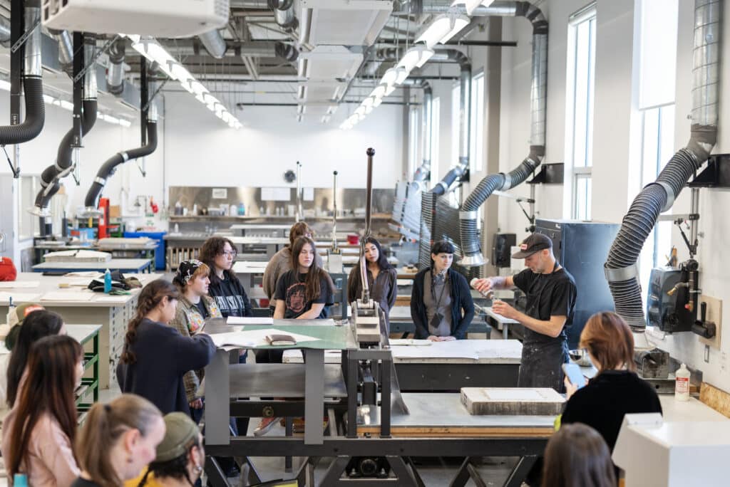 A group of students listen to an instructor demonstrating in a printmaking studio with large equipment.