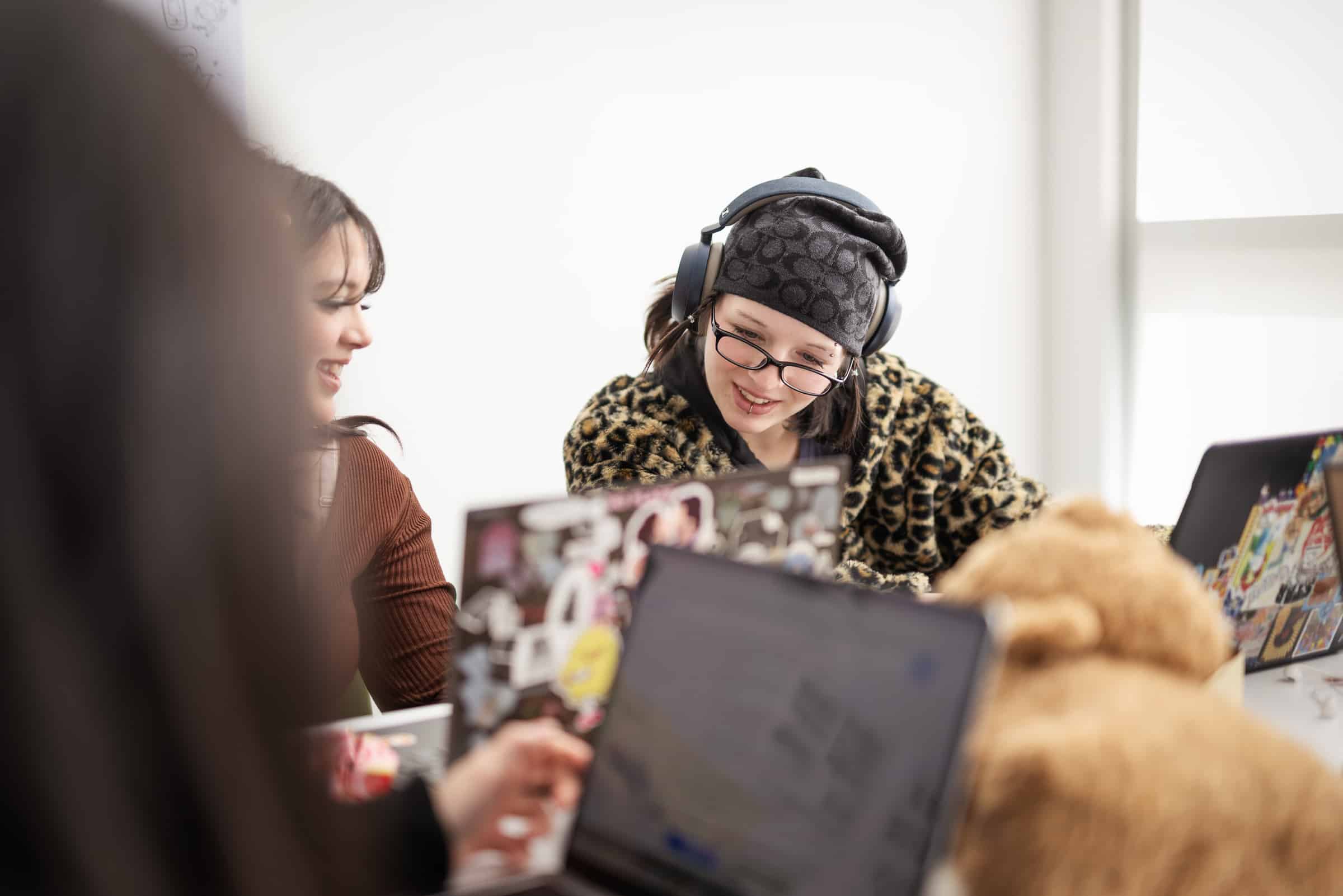 Two students work together at laptops, smiling and talking in a classroom setting.
