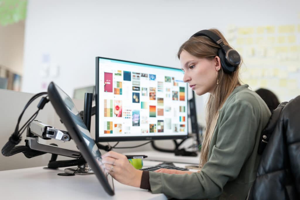 A close-up of a student wearing headphones and using a stylus on a drawing tablet, while a large monitor in front of her displays a grid of colorful images. She works intently in a modern computer lab.