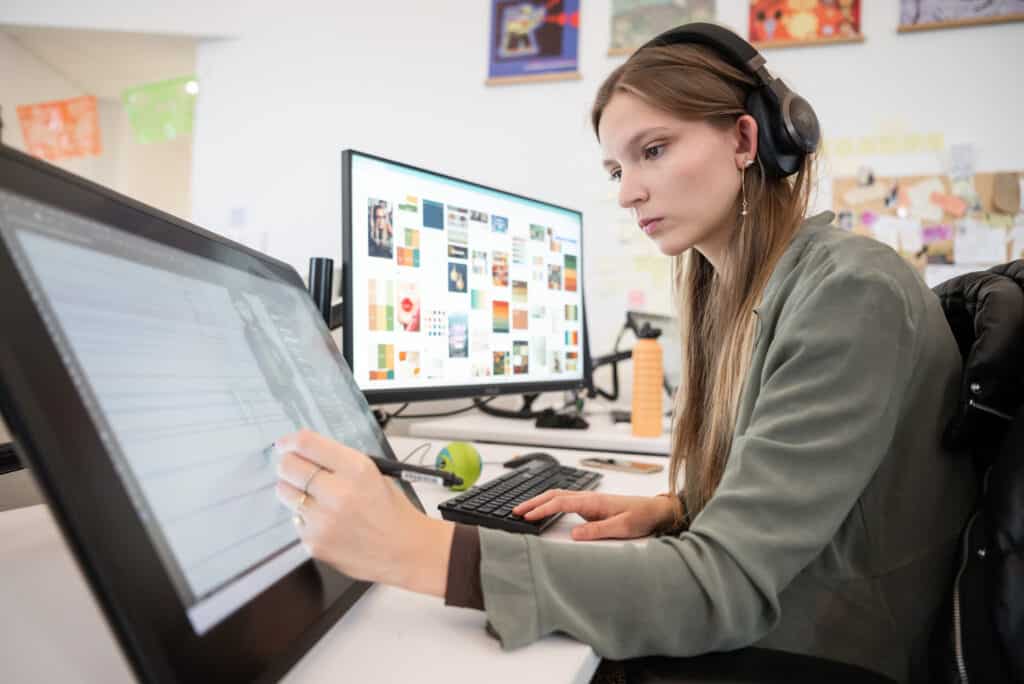 Person wearing headphones working on a digital drawing tablet at a desk, with a large monitor in the background displaying a color reference board.