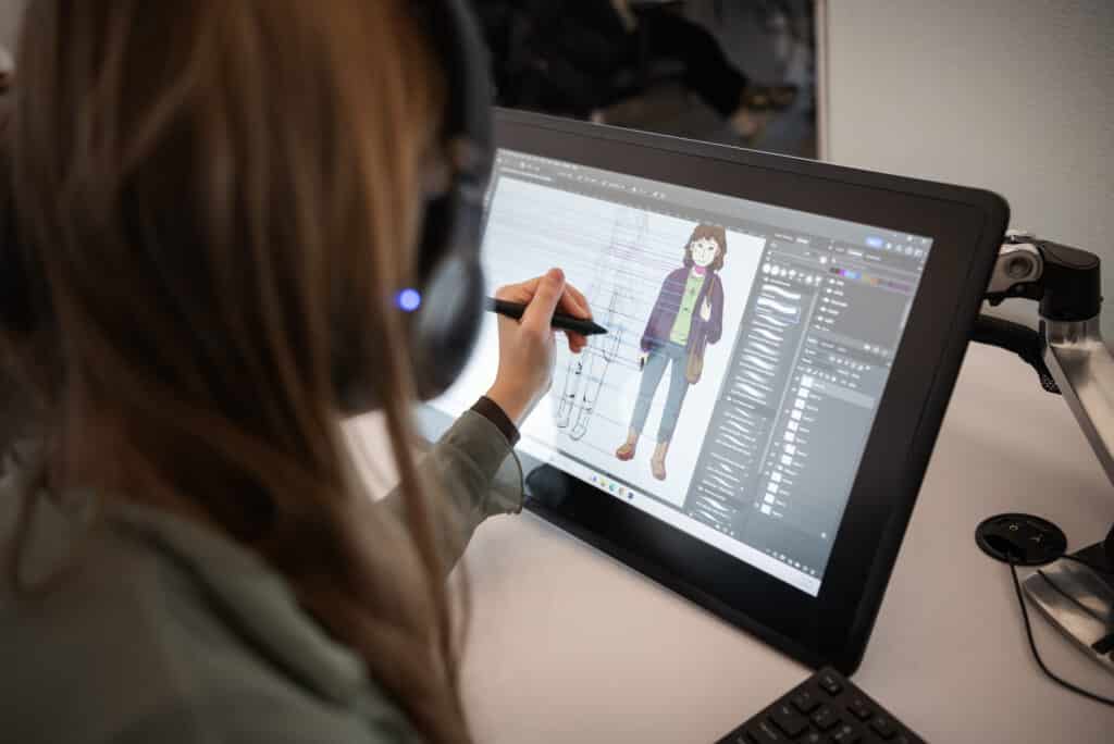 A person wearing headphones draws digitally on a pen display tablet, creating a colorful character illustration of a person in casual clothes using design software.