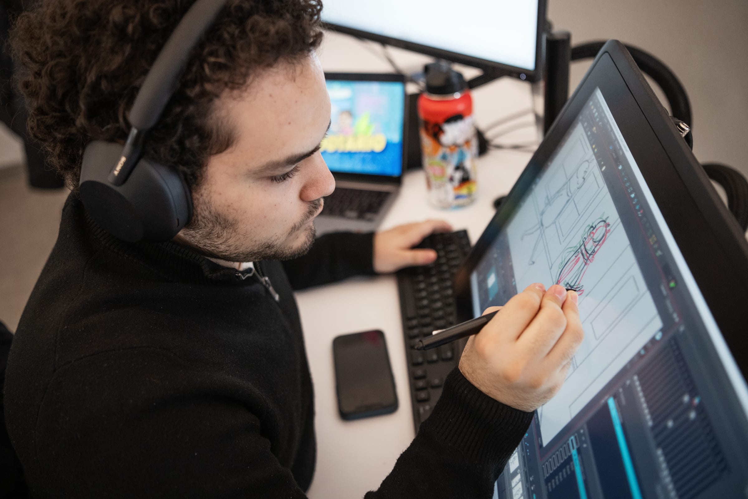 Person wearing headphones using a pen tablet to draw storyboards on a screen, with a keyboard, water bottle, and small monitor in the background.