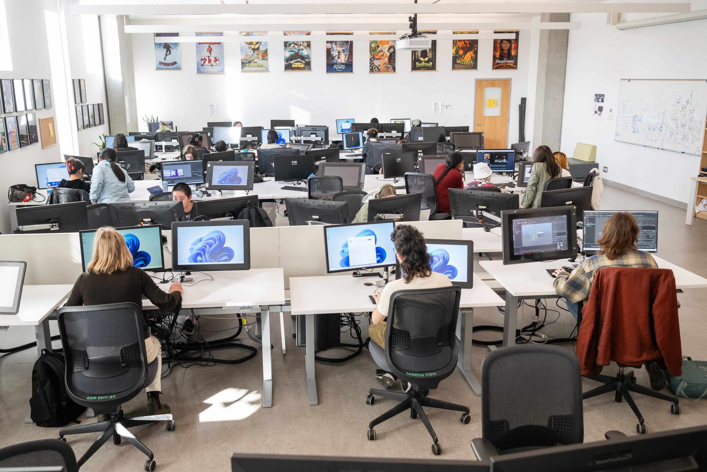 A wide shot of a computer lab where many students work at large monitors and drawing tablets. The room is bright, with posters of animated films on the walls. Students focus on digital projects in a collaborative classroom environment.