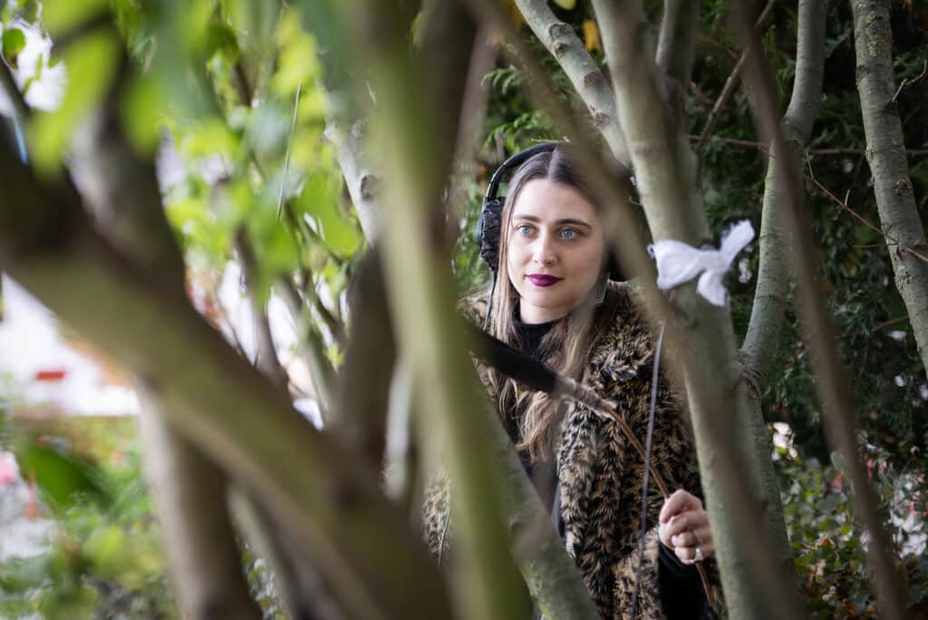 A woman wearing headphones and a faux-fur coat stands among tree branches, holding a stick with a white ribbon tied to it. She looks focused, with bright lipstick and blue eyes, framed by greenery in the background.