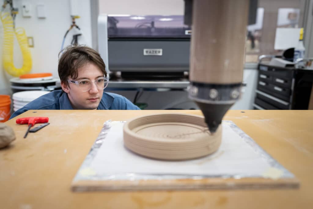 Person wearing safety glasses closely watches a 3D clay printer as it builds a circular layered form on a flat surface. Tools and equipment are visible in the workshop background.