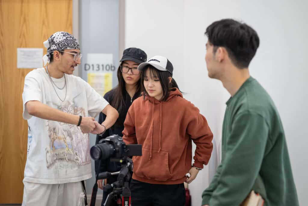 A group of four students gathered around a camera on a tripod in a classroom, with one student gesturing while explaining something to the others.