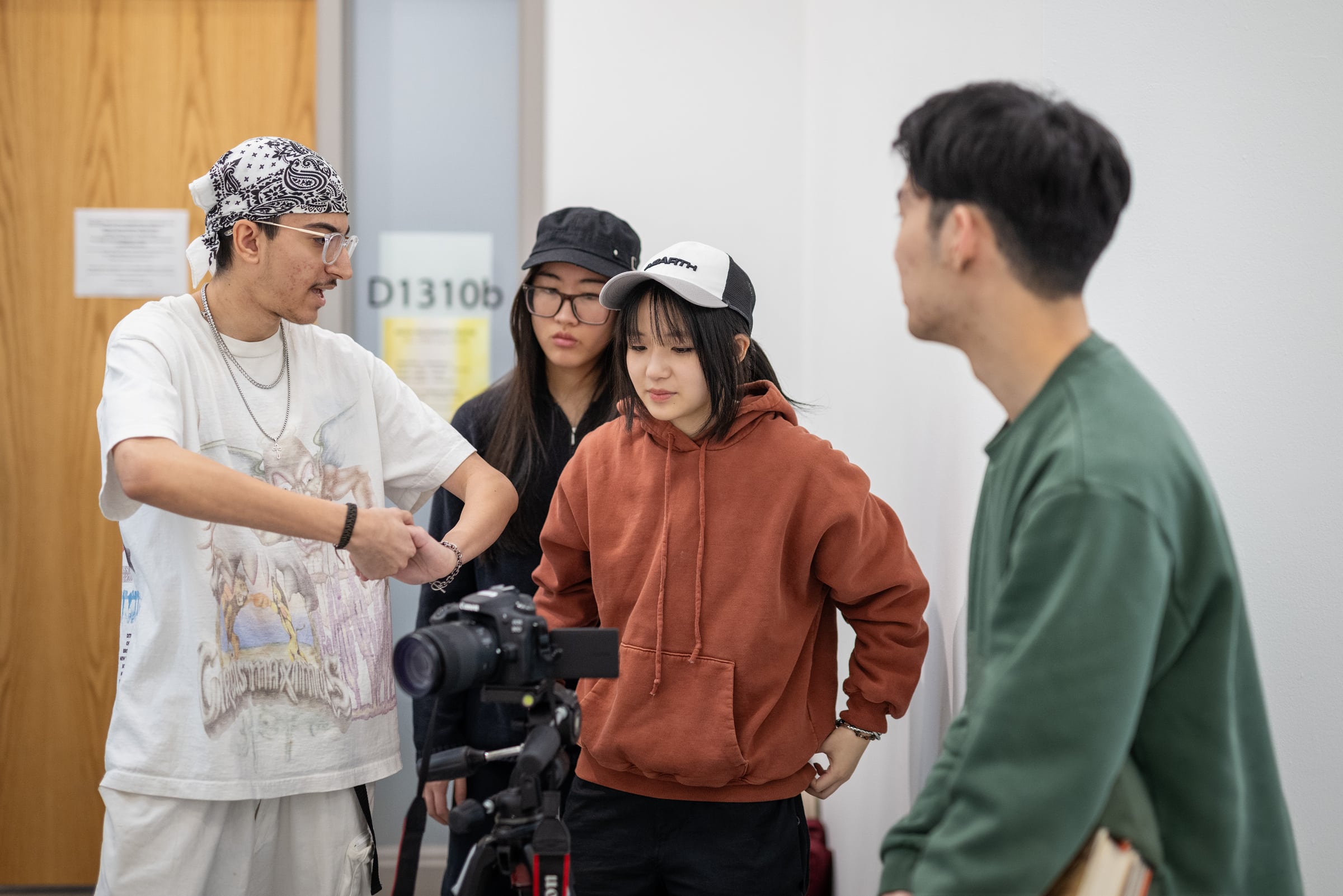 A group of four students gathered around a camera on a tripod in a classroom, with one student gesturing while explaining something to the others.