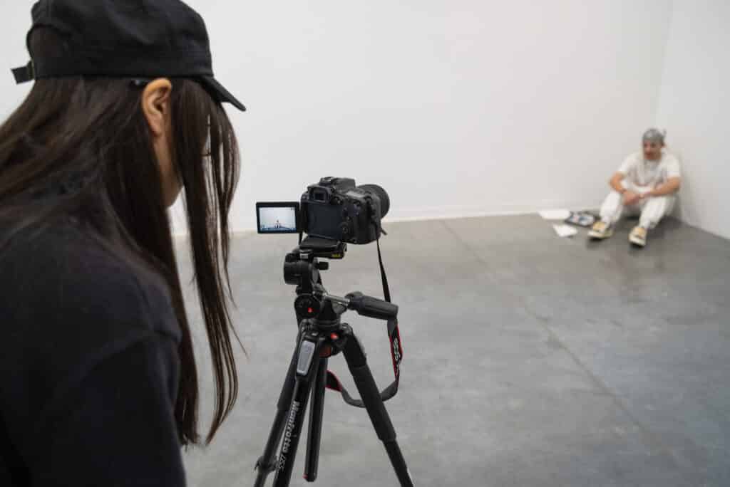 A student in the foreground with her back towards us looks through the viewfinder of a camera. Another student, several feet away, sits on the floor in a corner of the room and is framed in the camera's viewfinder.