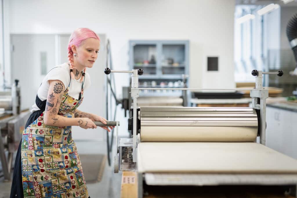 Person with pink hair in braids operates a large printmaking press in a studio. They wear a patterned apron and turn the handle to run a print through the press.