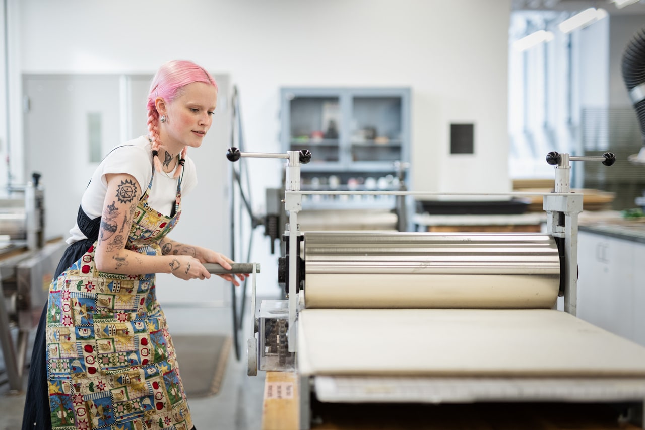 Person with pink hair in braids operates a large printmaking press in a studio. They wear a patterned apron and turn the handle to run a print through the press.
