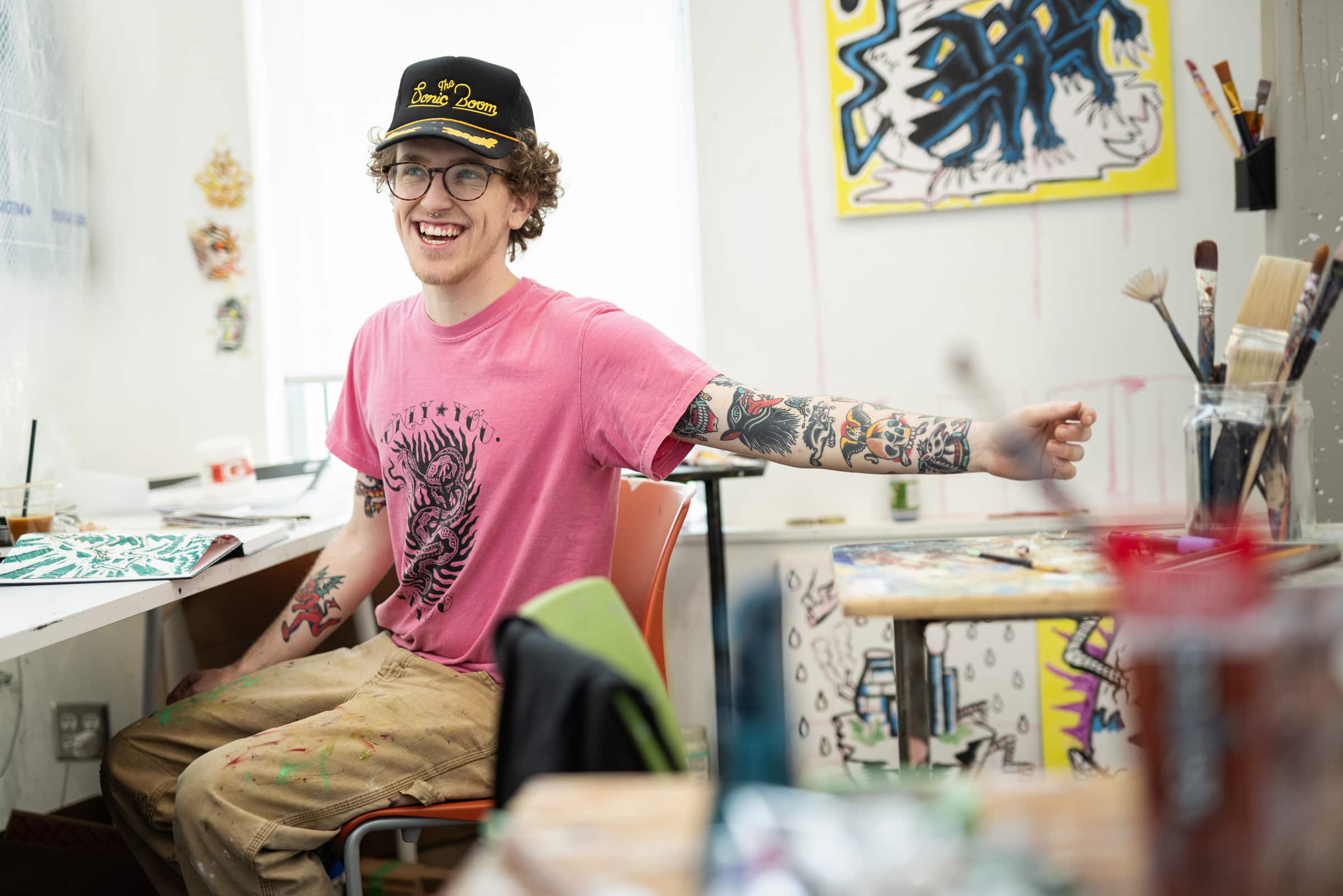 Person in a pink shirt and cap sits at a paint-splattered desk, smiling and extending their tattooed arm. Art supplies and colourful paintings surround the workspace.