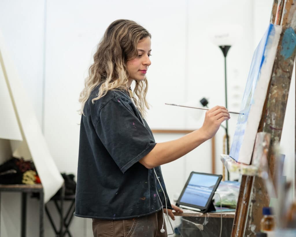 Student painting on a canvas at an easel in a studio, holding a brush while referencing an image on a tablet nearby.