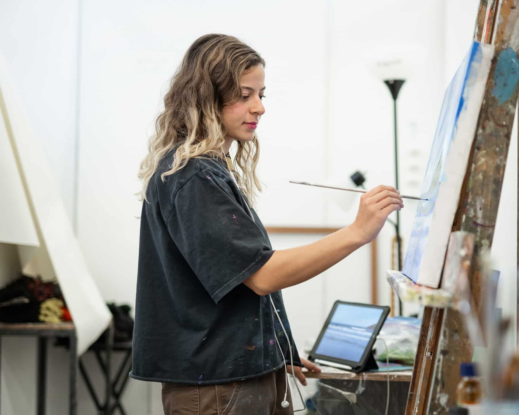Student painting on a canvas at an easel in a studio, holding a brush while referencing an image on a tablet nearby.
