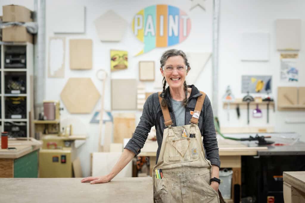 Person in a workshop wearing safety glasses and overalls stands smiling at a workbench. The background wall is covered with geometric wooden panels, tools and a colourful sign that reads “PAINT.”