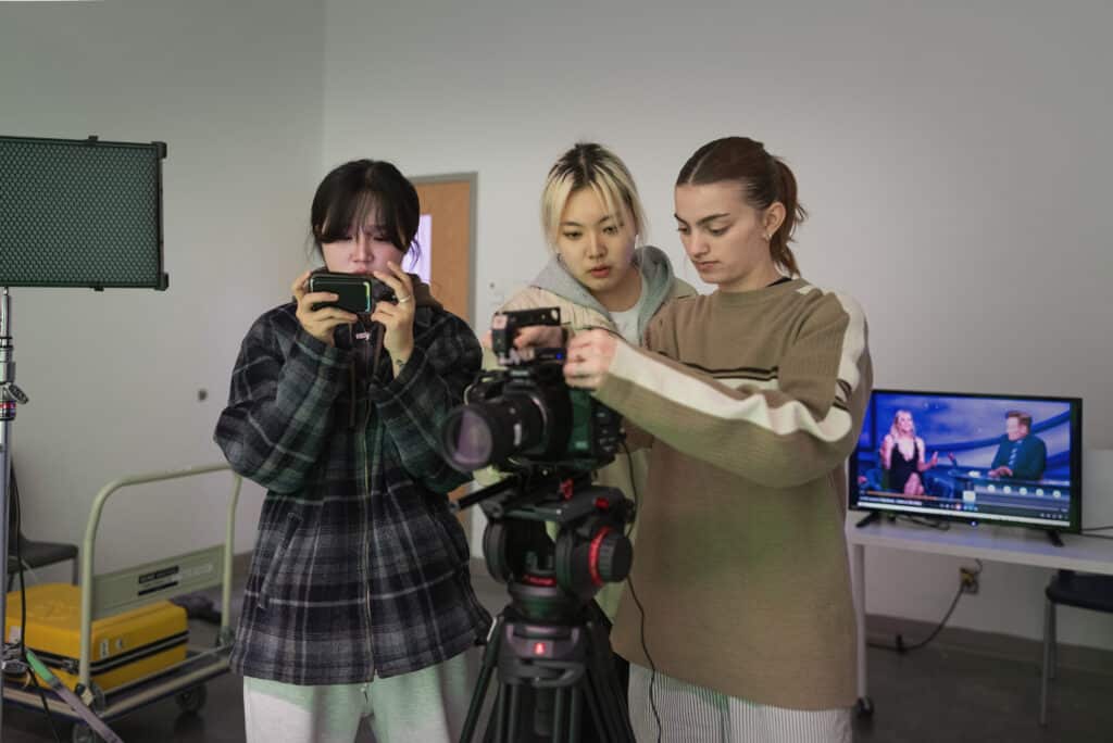 Three students setting up a professional video camera in a studio, with one adjusting the camera, another holding a phone, and a TV screen in the background.
