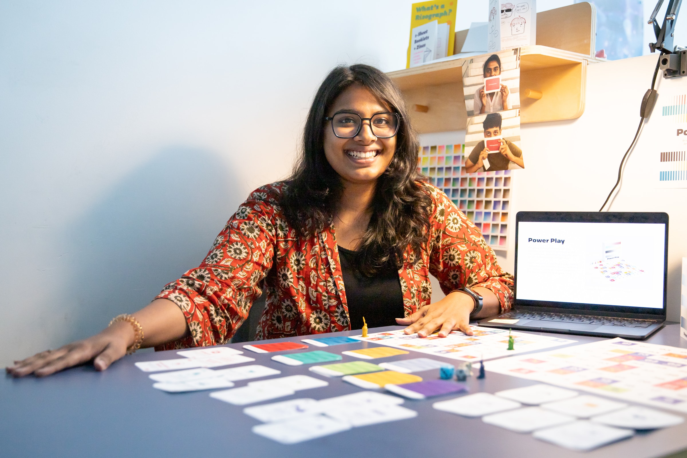 A smiling student sits at a desk with a colourful board game prototype laid out in front of them, with a laptop displaying the project.