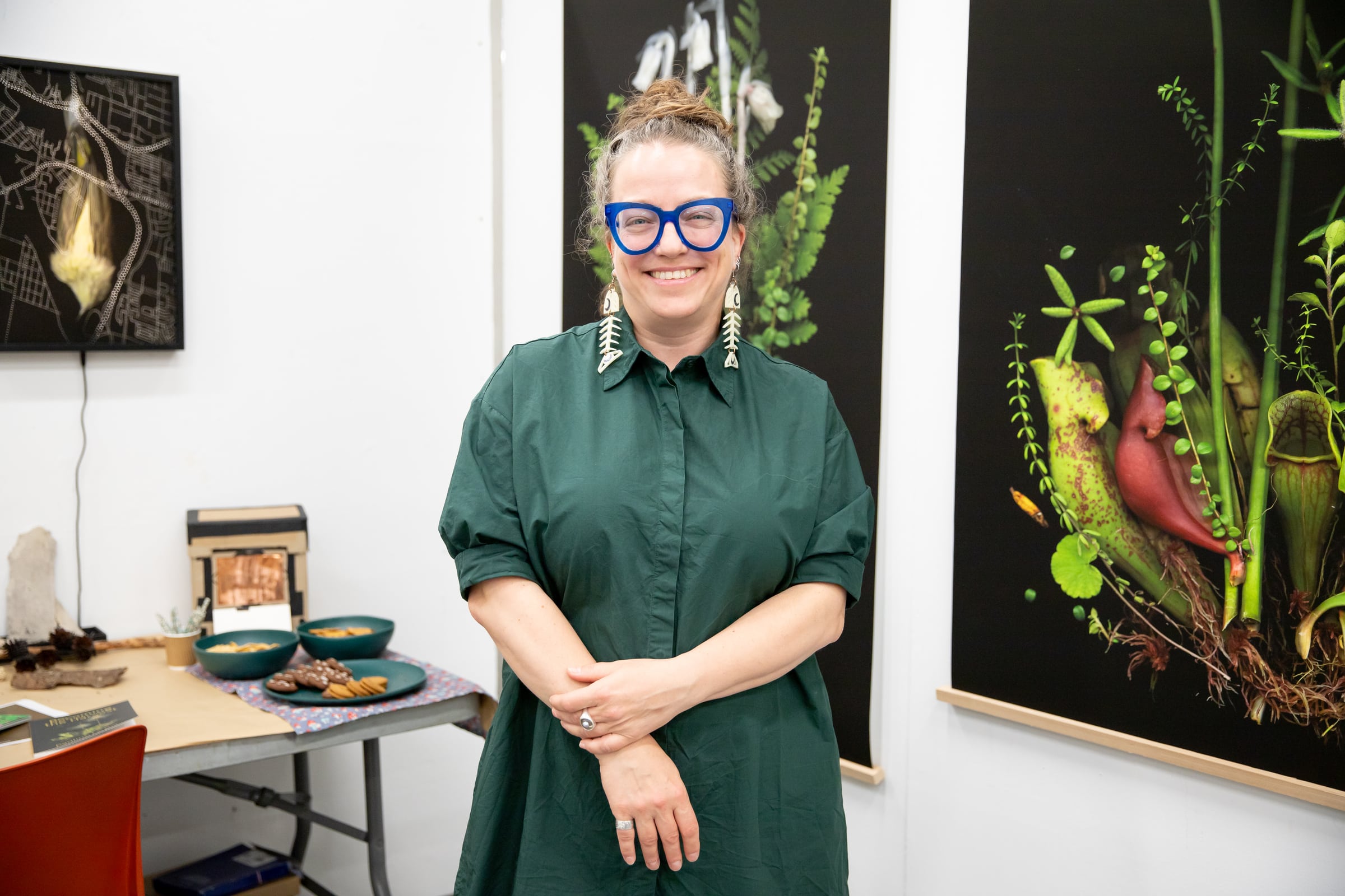A smiling person wearing a green dress and large blue glasses stands in front of botanical artworks in a studio or gallery.