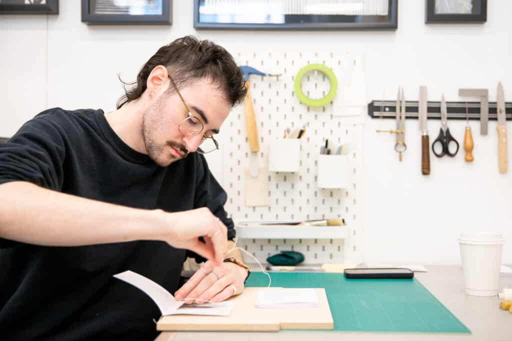 A student with short dark hair and glasses sews pages together while bookbinding at a work table, with tools hanging neatly on the wall behind them.