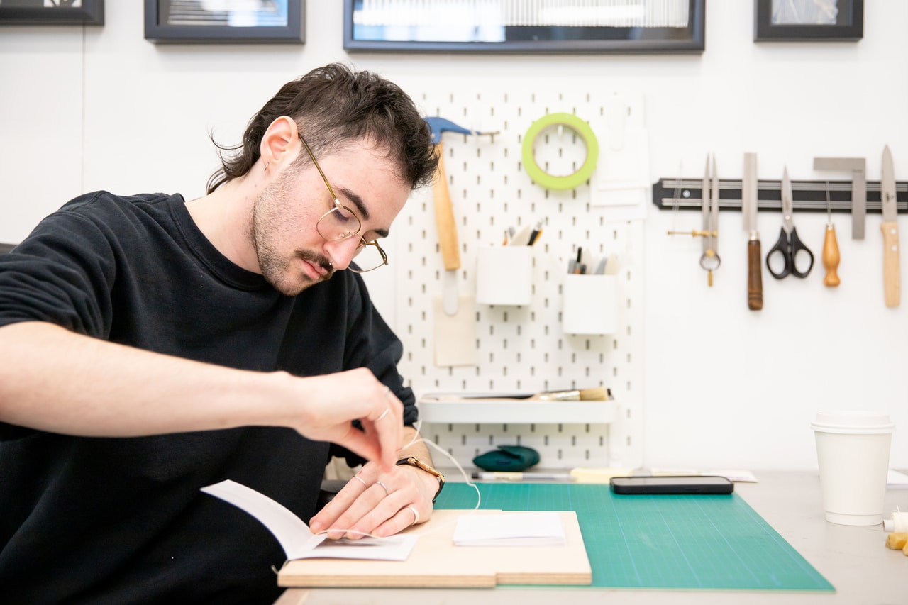 A student with short dark hair and glasses sews pages together while bookbinding at a work table, with tools hanging neatly on the wall behind them.