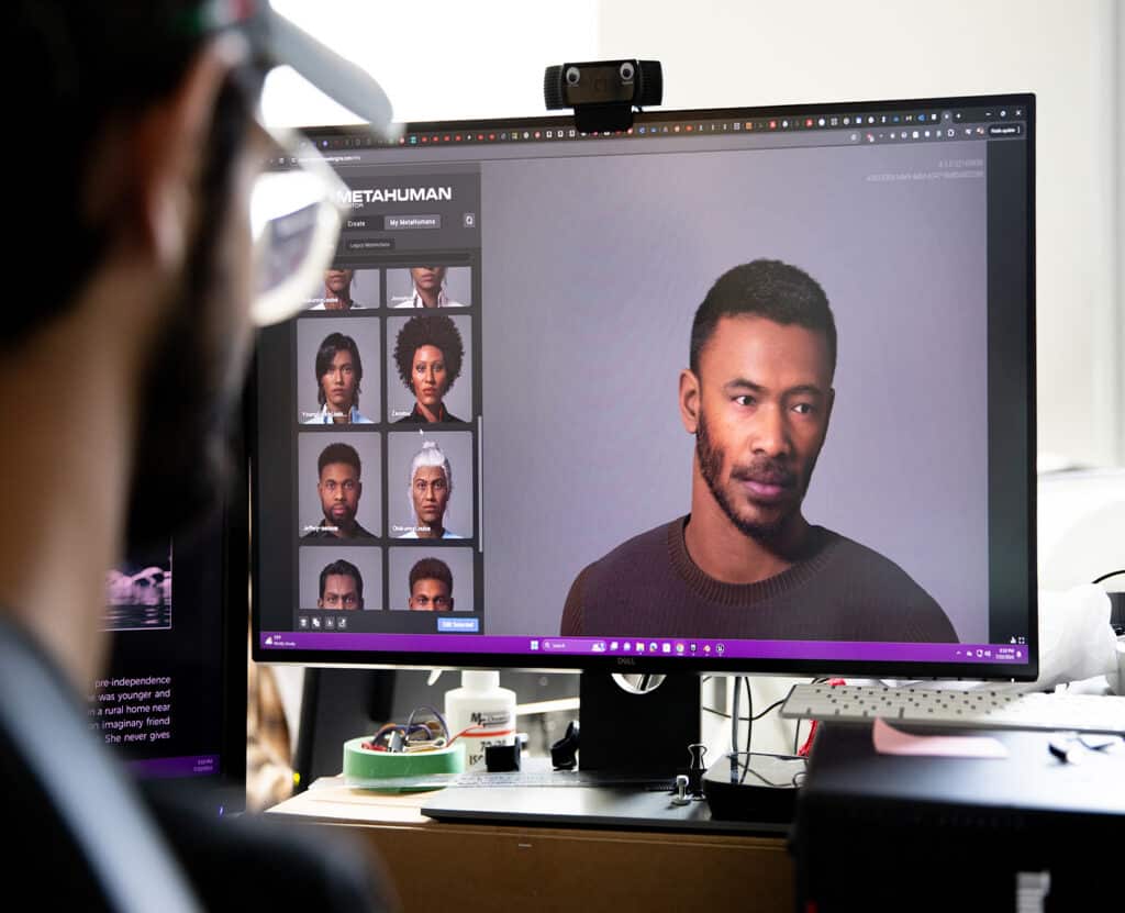 A person wearing glasses looks at a computer screen displaying Epic Games’ MetaHuman Creator. The monitor shows several digital character options on the left and a realistic 3D-rendered man with short hair and a beard on the right.
