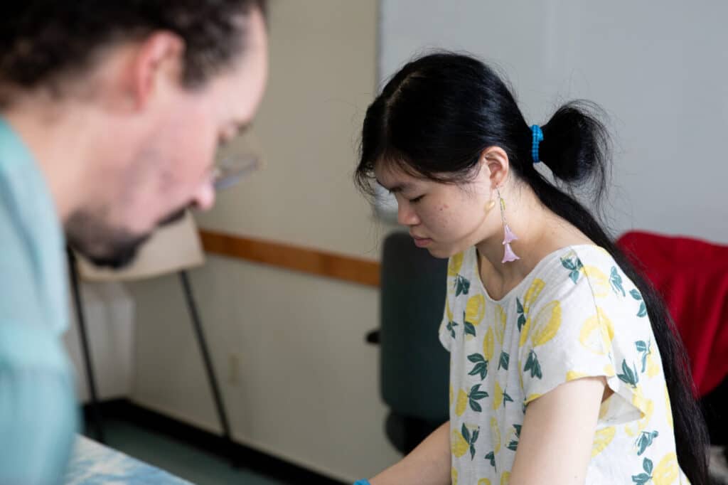 A person in a patterned shirt works intently at a table, with another person blurred in the foreground.