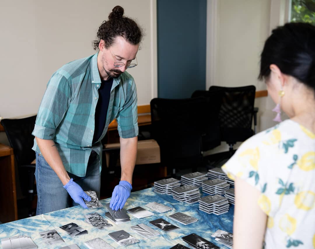 A person wearing a green plaid shirt, jeans, and blue gloves sorts through small black-and-white printed cards laid out on a table covered with a blue patterned cloth. Stacks of cards are neatly arranged on the table, while another person in a lemon-patterned shirt observes from the side. The setting appears to be an indoor workspace with chairs and desks in the background.