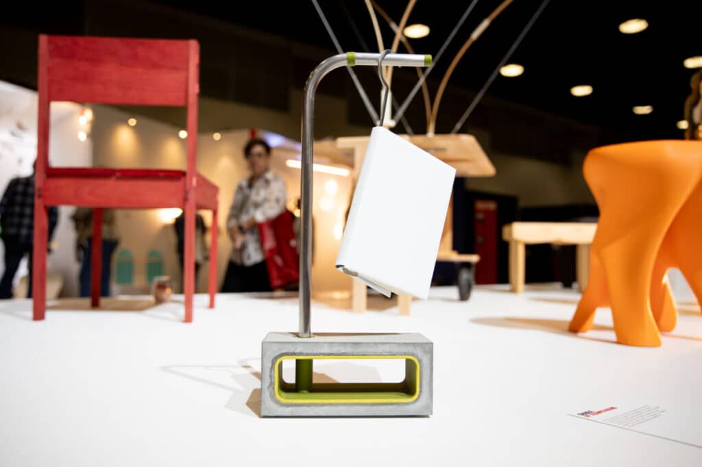 A minimalist lamp with a rectangular concrete base and a curved metal rod holding a white shade, displayed on a pedestal with other contemporary furniture pieces in the background.