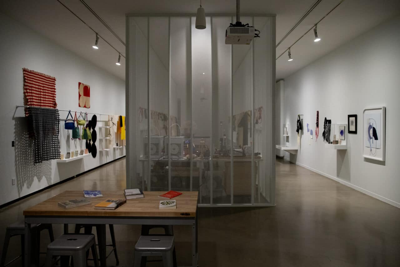 Gallery view showing textiles, garments, and accessories displayed on walls and shelves around a central enclosed studio space.