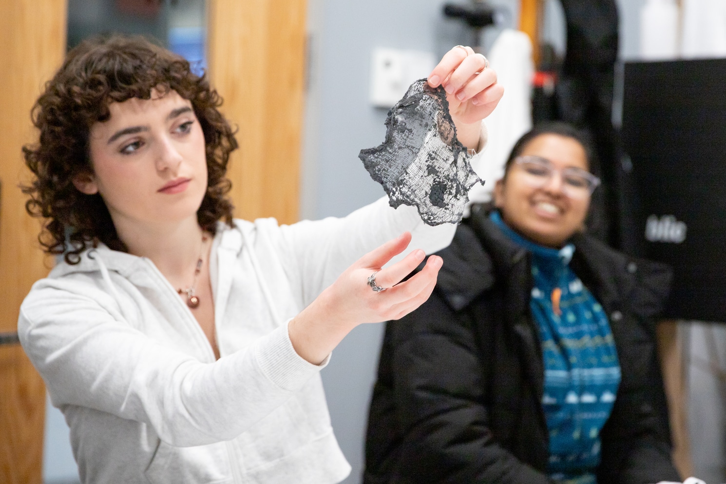 A student holds up a thin, charred black material sample for examination while another person in the background smiles during a classroom discussion.