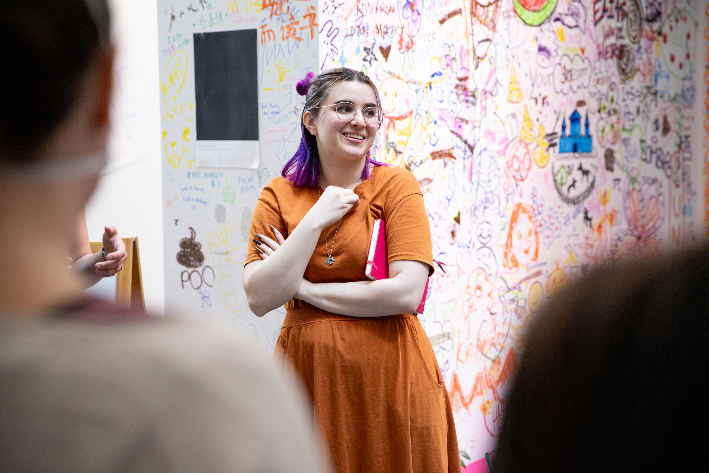 Student in orange dress leads a discussion in front of a wall covered in colorful doodles, text, and sketches, holding a pink notebook.