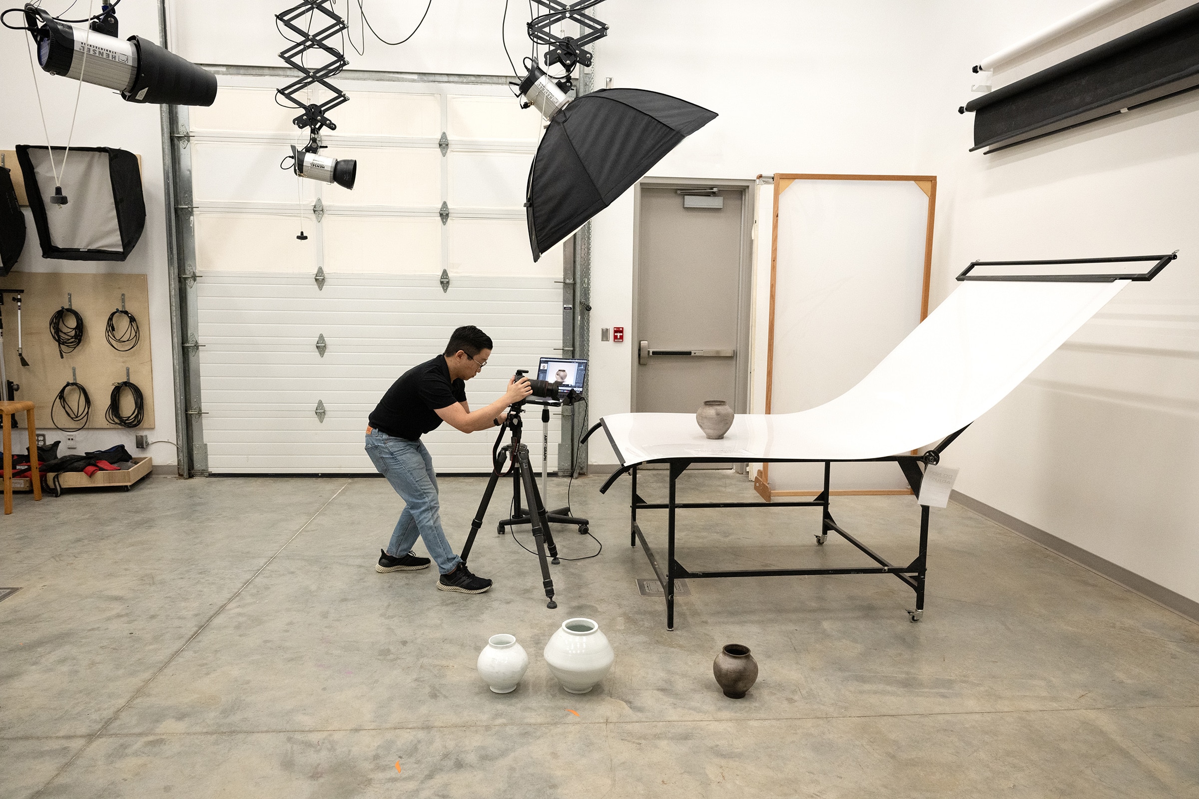 A person in a photography studio taking pictures of ceramic vases on a white backdrop, surrounded by studio lights and equipment.