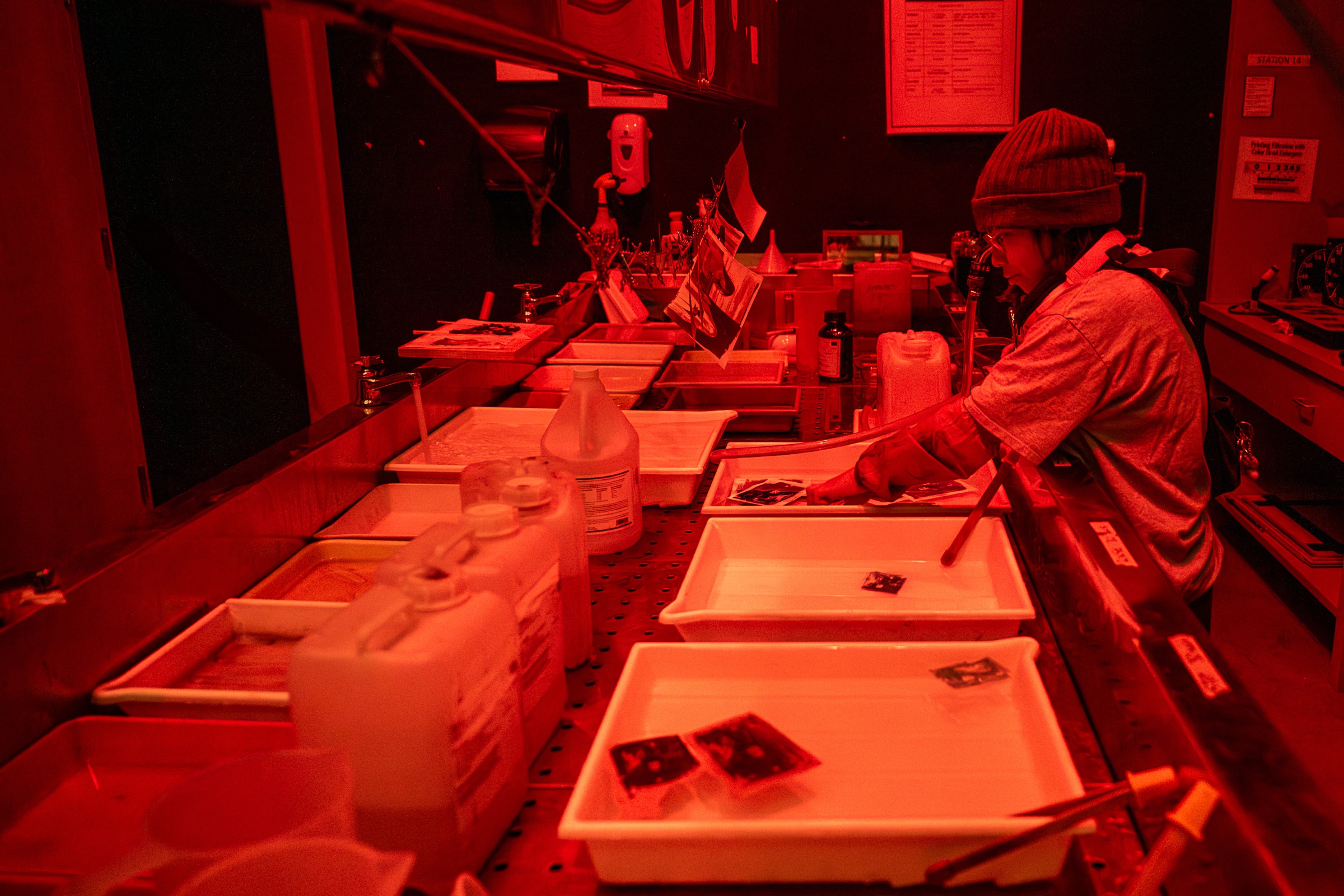 A person in a darkroom working with film prints under red lighting, using chemical trays for developing.