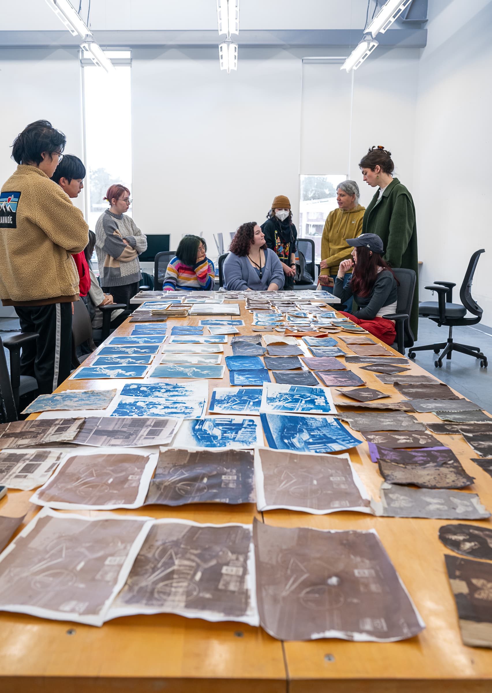 A group of students standing around a large table covered with blue and brown-toned photographic prints, engaged in discussion.