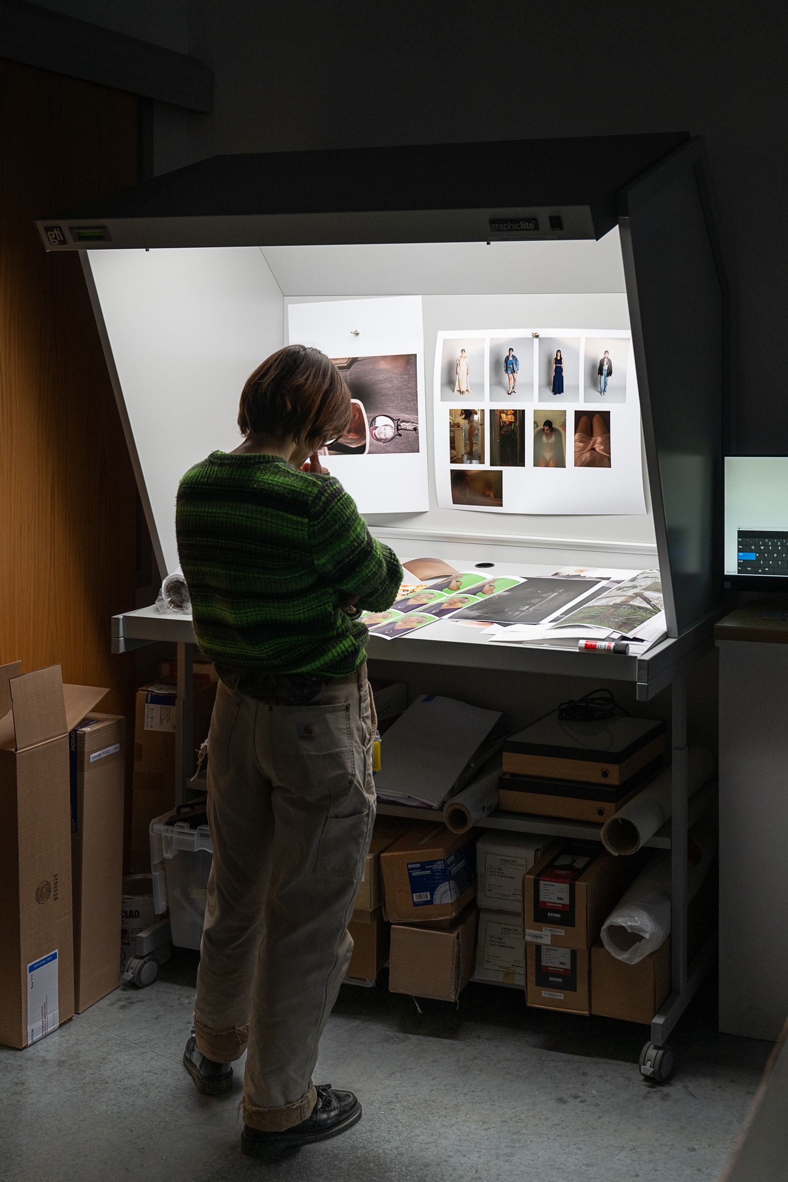 A person standing at a light table reviewing several photographic prints pinned to the wall under bright inspection lighting.