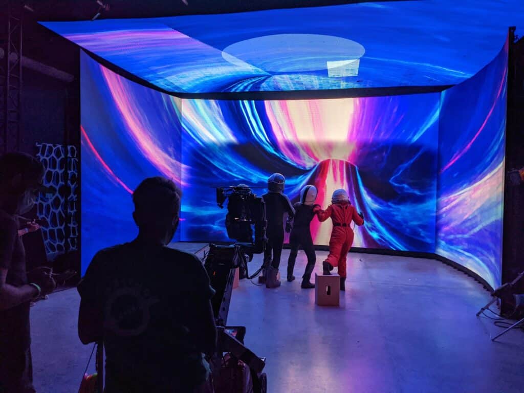 A behind-the-scenes view of a virtual production shoot inside Emily Carr University’s Integrated Motion Studio. Three performers in futuristic space suits stand before a massive, curved LED volume displaying a vibrant, swirling cosmic tunnel in blues, pinks, and yellows. A film crew operates a camera rig nearby, capturing the scene against the immersive digital backdrop — showcasing how physical sets are replaced by computer-generated environments.