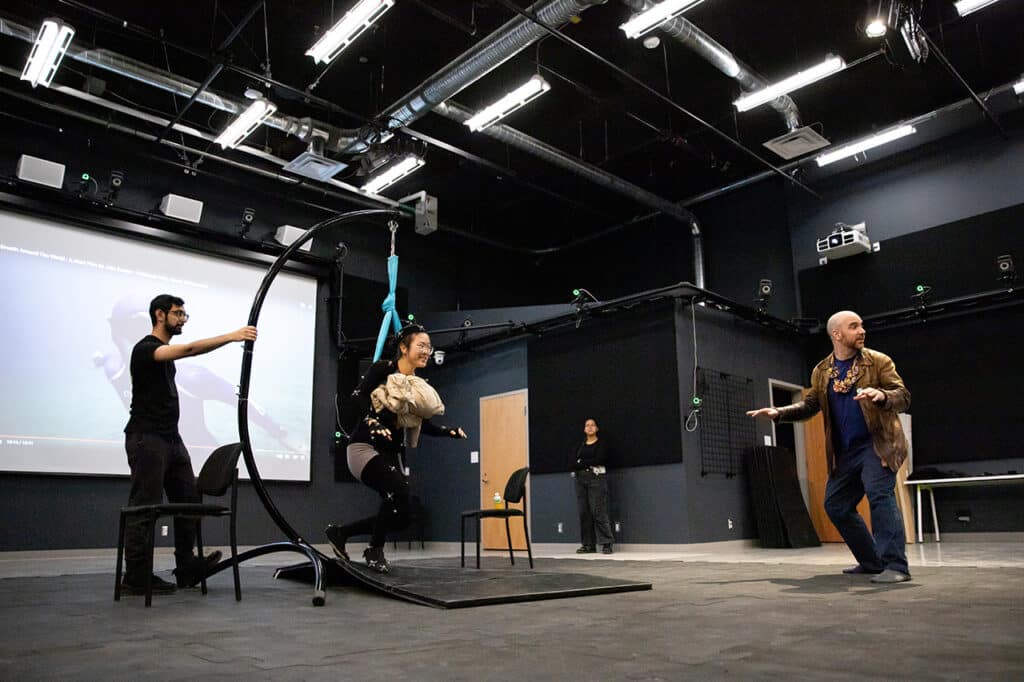 Motion capture studio with black walls and overhead lighting. A performer in a harness and mocap suit is lifted while interacting with a director, as crew members assist and observe.