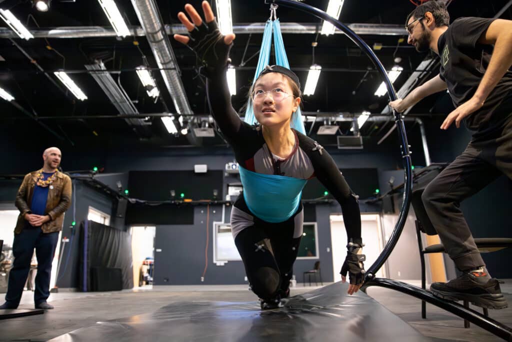 Student in a motion capture suit suspended in a harness, reaching forward while instructors observe and assist in a studio equipped with sensors and lights.