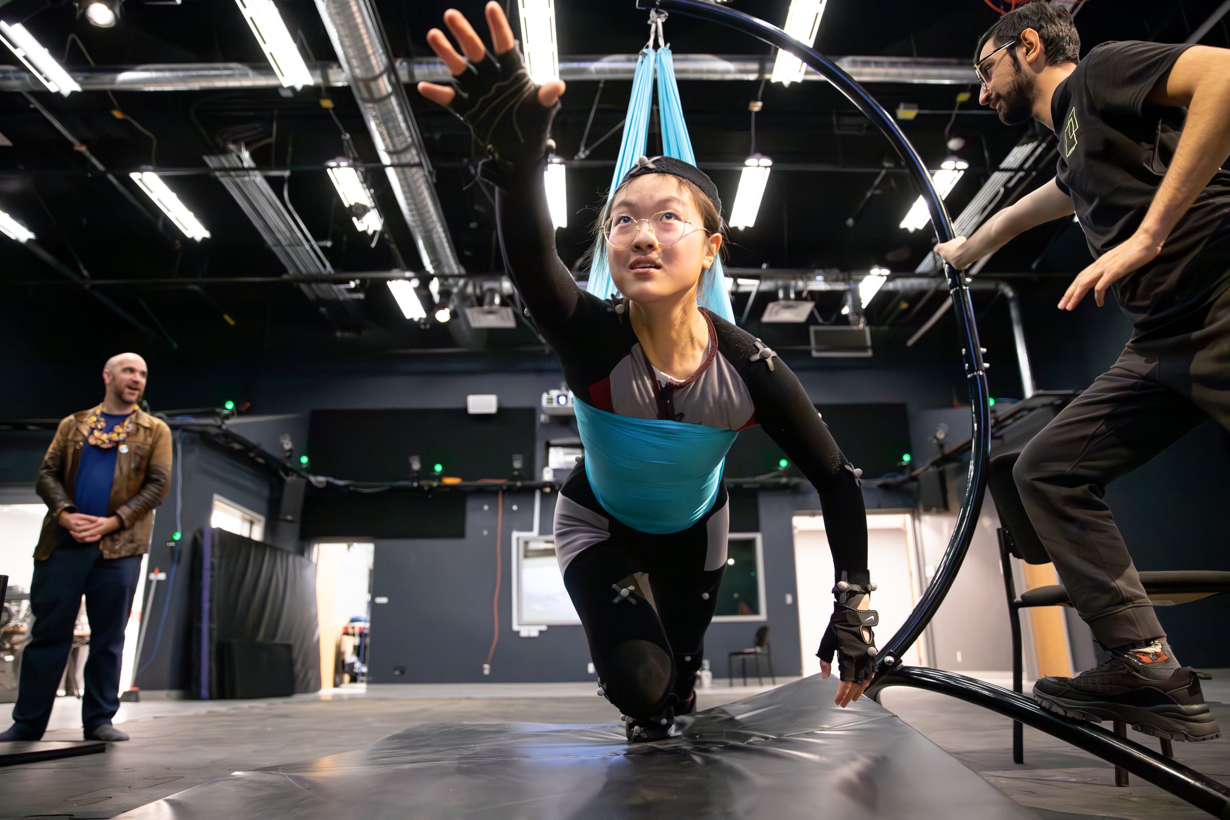 Student in a motion capture suit suspended in a harness, reaching forward while instructors observe and assist in a studio equipped with sensors and lights.