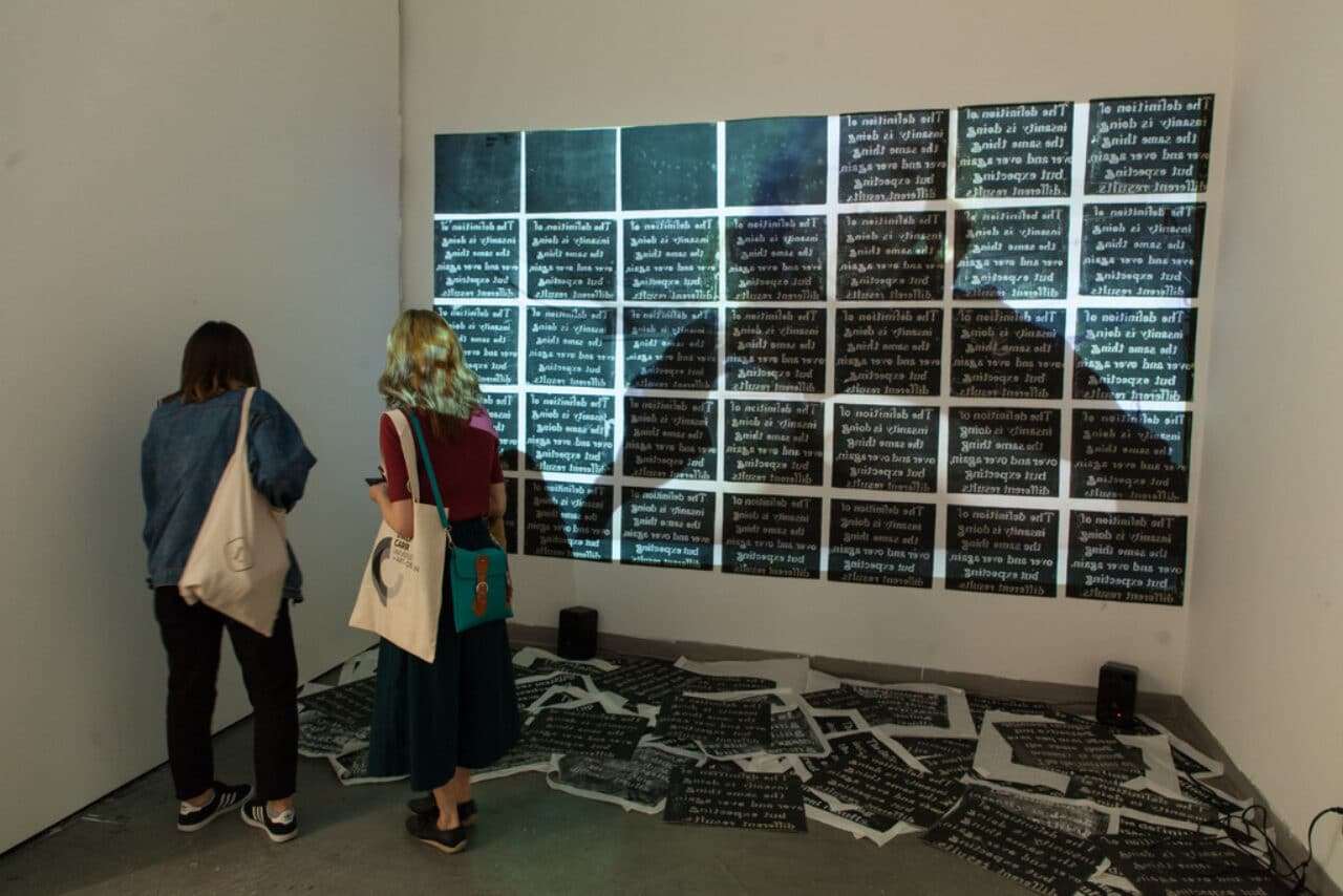 Two visitors stand in front of a wall projection consisting of repeated squares of black-and-white text. Printed sheets with the same text are scattered on the floor, creating an immersive installation.
