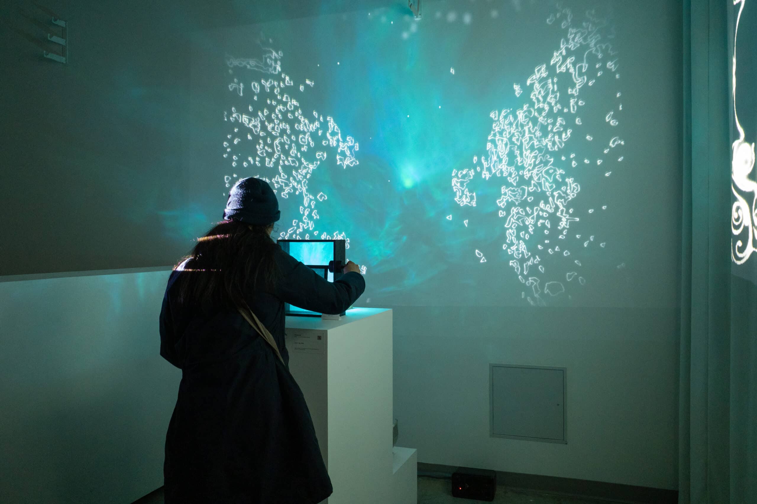 A guest, seen from behind wearing a dark coat and beanie, interacts with Sienna Lee’s 2025 installation “Close-Up” at Emily Carr University’s Show 2025. The guest leans over a white pedestal to adjust a small device projecting an ethereal, turquoise-hued video onto the wall. The projection features delicate, glowing white line drawings resembling abstract organic forms or constellations, floating against a soft, misty background.