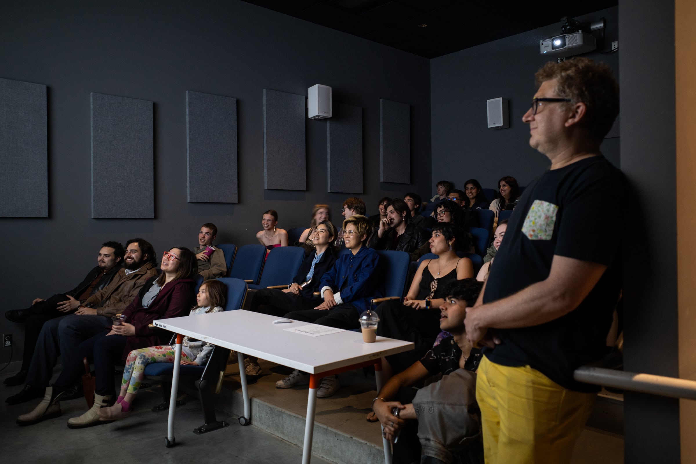 An audience of students and a professor watching a film screening in a small theater, smiling and engaged.