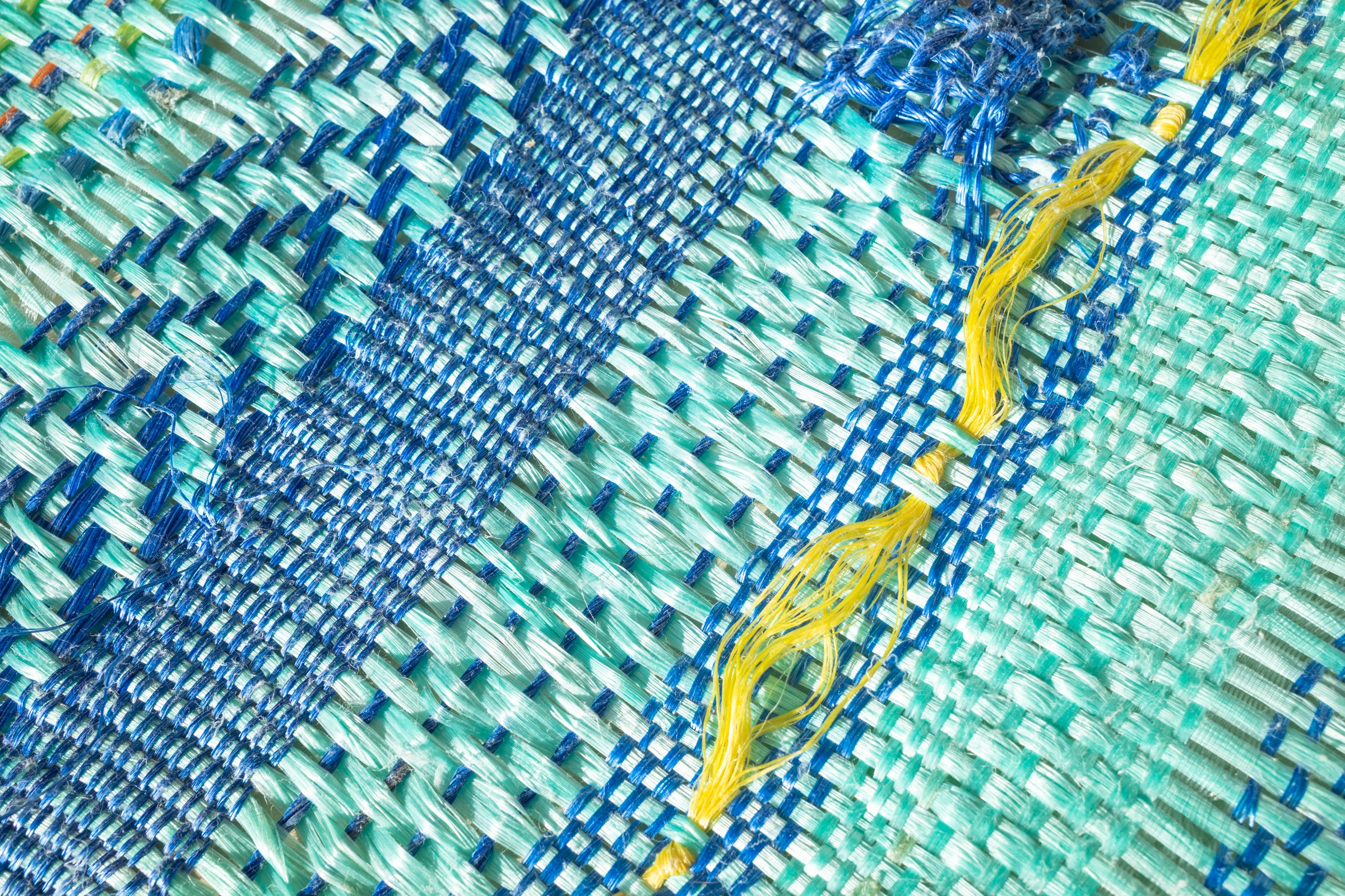 Detail of a woven textile in turquoise and blue with yellow threads, showing texture and interlaced fibers up close.